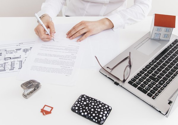 A hand signing property paperwork beside a laptop and a floor plan